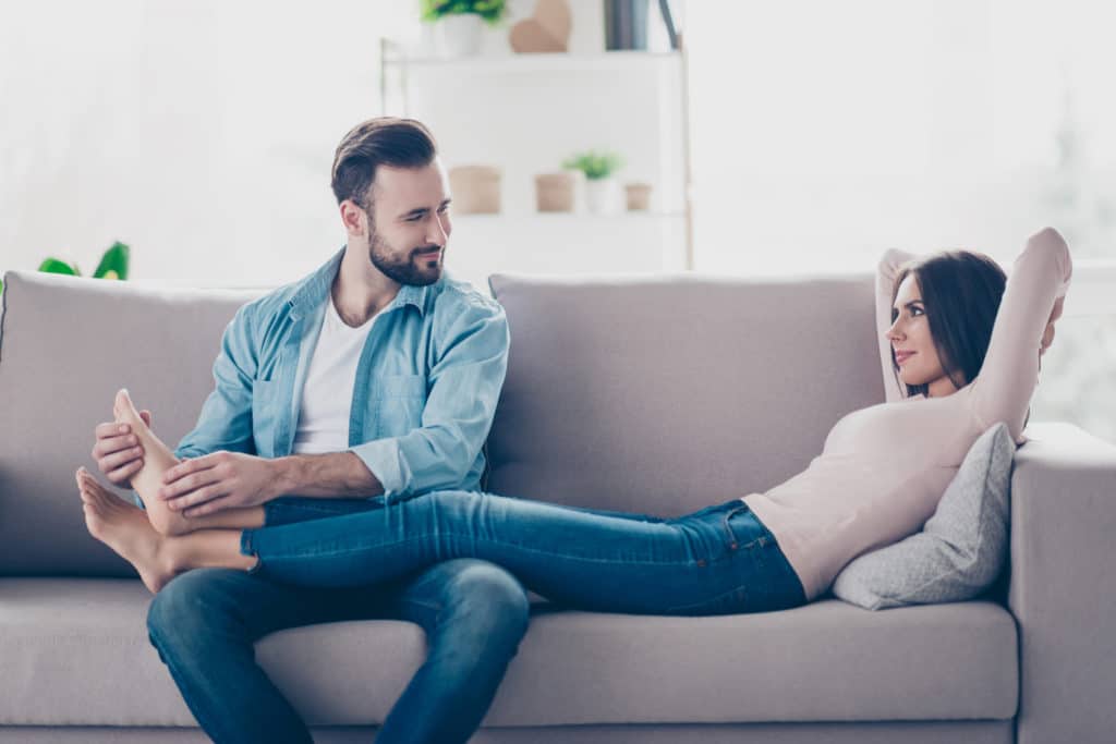 Attentive caring loving handsome bearded husband is doing feet massage for her attractive tender lying on a sofa wife, work at it